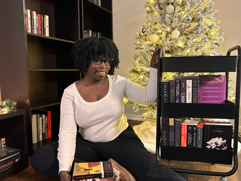 photo of Kathleen sitting by her bookshelf touching her TBR cart with a stack of books on the floor and a christmas tree in the background showing off her winter tbr 2024