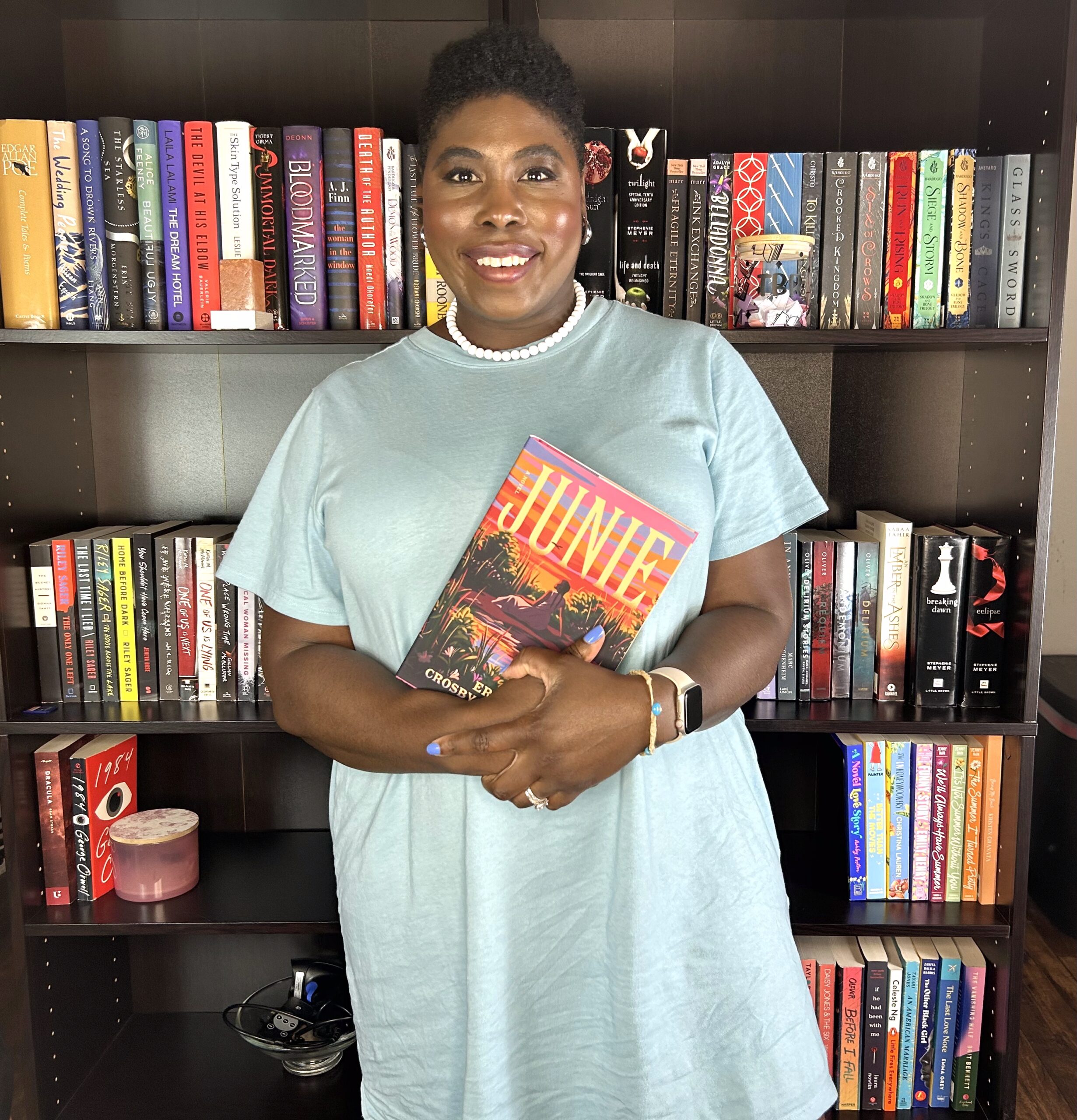 photo of kathleen haagenson holding her favorite book of June standing in front of her bookshelves